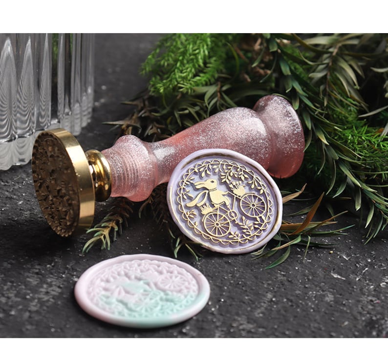 Pu&ograve; includere: Un timbro per sigilli in cera rosa e oro con un manico decorativo e un disegno di un coniglio che guida una bicicletta. Il timbro &egrave; su una superficie grigio scuro con vegetazione verde.