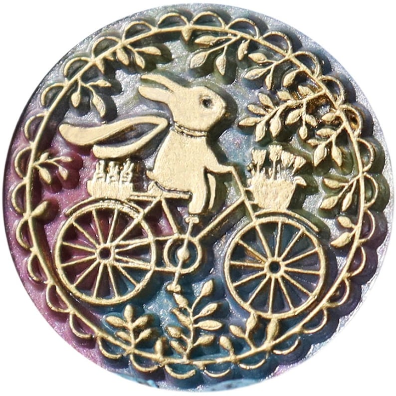 Pu&ograve; includere: Un ciondolo in metallo dorato con un design dettagliato di un coniglio che guida una bicicletta con un cestino di fiori. Il ciondolo &egrave; circondato da un bordo floreale decorativo e ha uno sfondo iridescente.