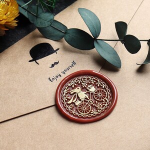 Pu&ograve; includere: Un sigillo di cera rossa con un disegno dorato di un coniglio che guida una bicicletta. Il testo "Enjoy yourself" &egrave; scritto su una busta marrone.