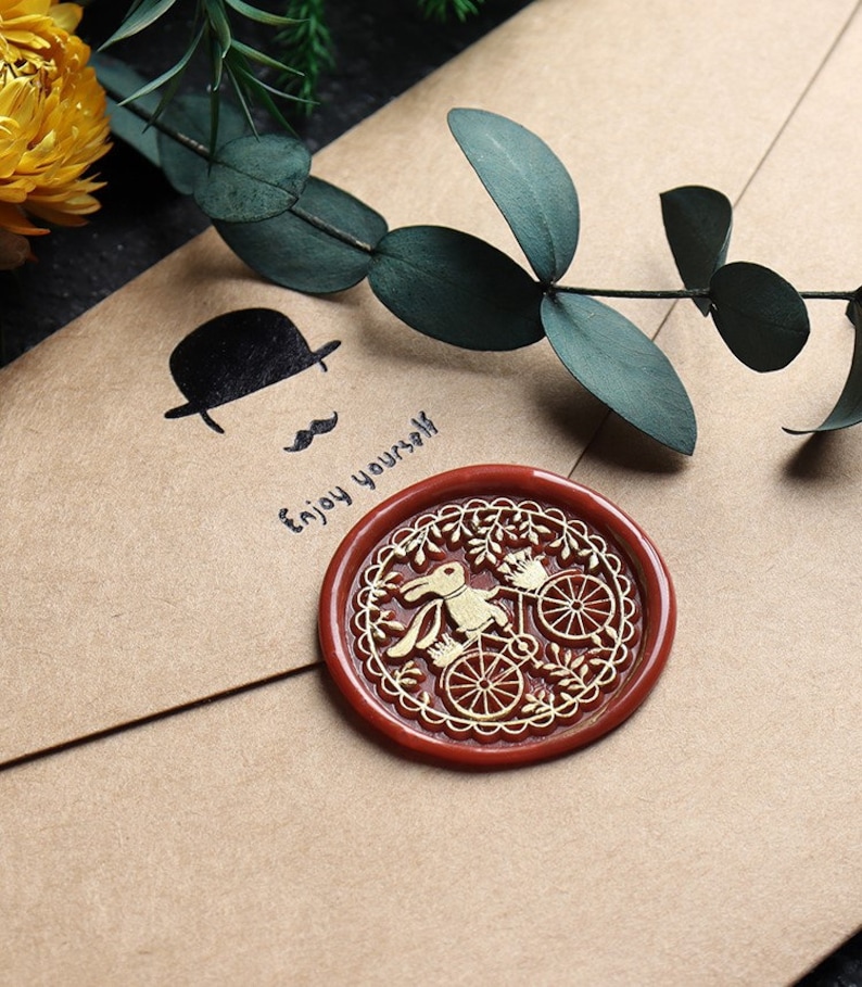 Pu&ograve; includere: Un sigillo di cera rossa con un disegno dorato di un coniglio che guida una bicicletta. Il testo "Enjoy yourself" &egrave; scritto su una busta marrone.