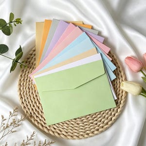 Pode incluir: Uma pilha de envelopes de papel retangulares em várias cores pastel, incluindo verde-claro, branco, azul-claro, rosa, lavanda, cinza, laranja e amarelo, dispostos em leque sobre um tapete de palha trançado redondo com textura lisa.