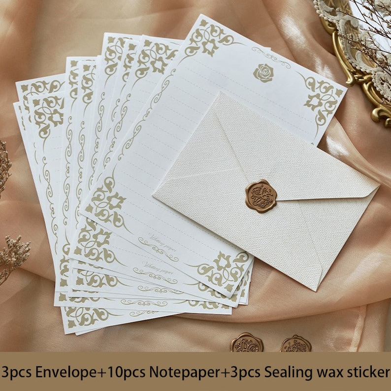 K&ouml;nnte beinhalten: Ein Set aus 10 wei&szlig;en Briefpapierbl&auml;ttern mit goldenen, verzierten R&auml;ndern und 3 wei&szlig;en Umschl&auml;gen mit einem goldenen Wachssiegel. Das Briefpapier hat Linien zum Schreiben und den Text "Writing paper" unten. Die Umschl&auml;ge haben eine strukturierte Oberfl&auml;che.