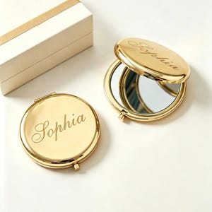 May include: Two round, gold-toned metal compact mirrors with a polished finish. One mirror is closed, and the other is open, revealing the reflective surface inside. Both feature the name Sophia engraved in a cursive script on the exterior lid.