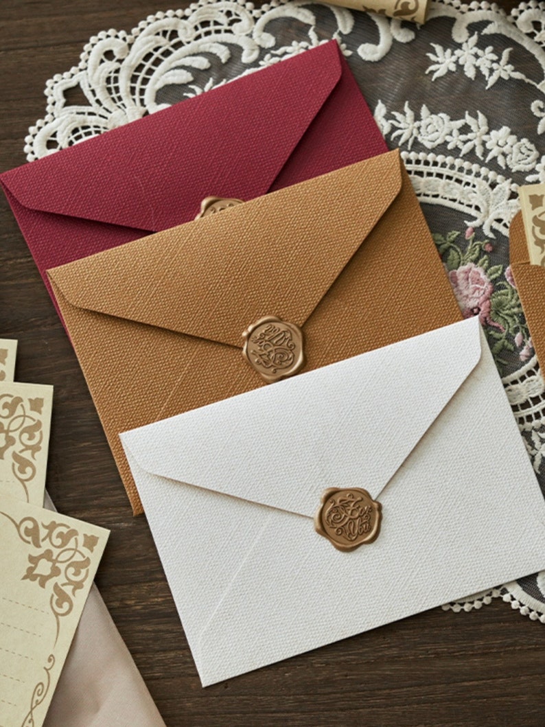 K&ouml;nnte beinhalten: Drei Umschl&auml;ge mit unterschiedlichem Papier und Wachssiegeln. Die Umschl&auml;ge sind braun, wei&szlig; und rot. Die Wachssiegel sind golden und haben ein Design.