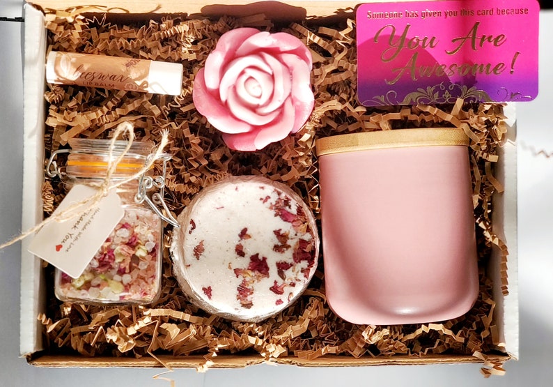 May include: A pink gift box filled with self-care items, including a pink rose soap, a jar of bath salts, a pink candle, and a lip balm. The box also includes a card that says "You Are Awesome!"