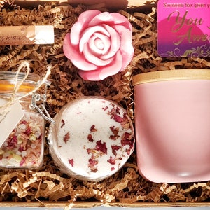 May include: A pink gift box filled with self-care items, including a pink rose soap, a jar of bath salts, a bath bomb, a lip balm, and a pink candle. The box also includes a card that says "You Are Awesome!"