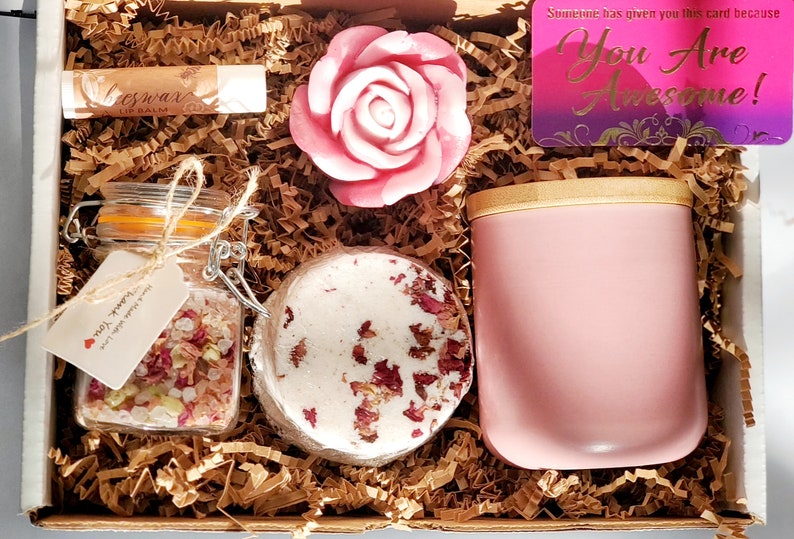 May include: A pink gift box filled with self-care items, including a pink rose soap, a jar of bath salts, a bath bomb, a lip balm, and a pink candle. The box also includes a card that says "You Are Awesome!"