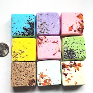 May include: Nine colorful, square bath bombs in various colors, including blue, purple, pink, yellow, green, brown, and white. Each bath bomb is decorated with dried herbs, seeds, or crystals. A quarter is next to the bath bombs for size comparison.