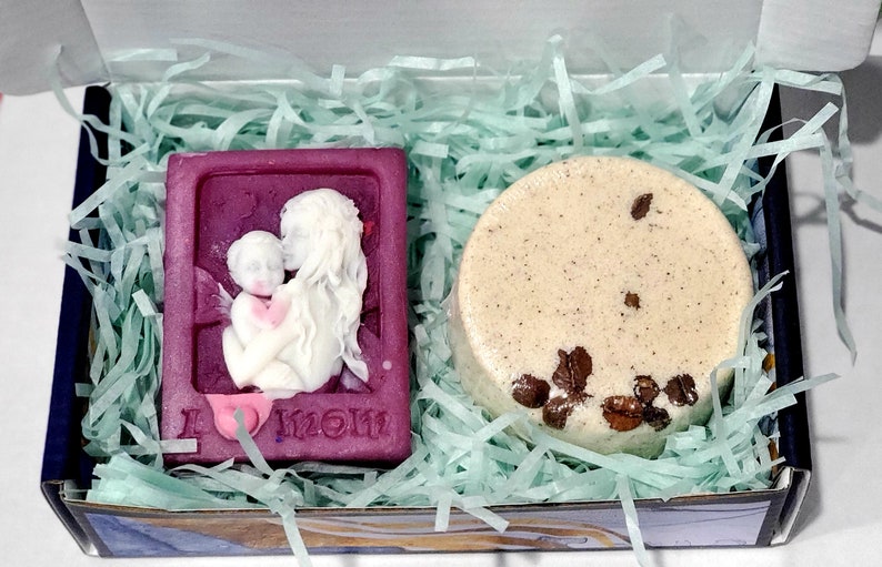 May include: A gift box filled with green shredded paper containing a purple soap bar with a white relief design of a mother and child and the text "I love mom" and a round beige soap bar with brown flecks.