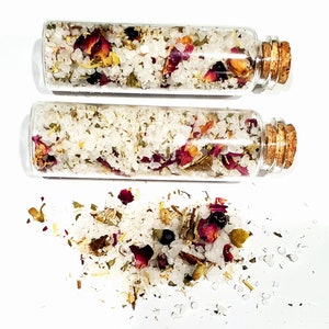 May include: Two small glass bottles filled with white bath salts, rose petals, and dried herbs. The bottles have cork stoppers and are sitting on a white surface. Some of the bath salts have spilled out of the bottles.