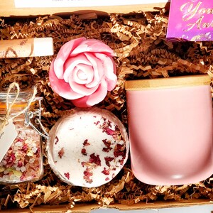 May include: A gift box filled with self-care items, including a pink rose-shaped soap, a bath bomb with rose petals, a jar of bath salts with rose petals, a pink candle with a wooden lid, and a lip balm. The box is lined with brown paper shred and includes a card that says "You Are Awesome!"