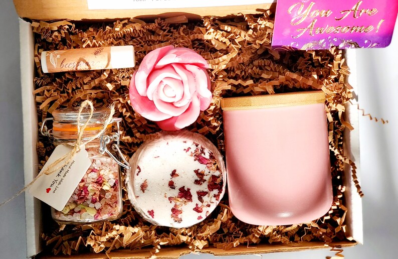 May include: A gift box filled with self-care items, including a pink rose-shaped soap, a bath bomb with rose petals, a jar of bath salts with rose petals, a pink candle with a wooden lid, and a lip balm. The box is lined with brown paper shred and includes a card that says "You Are Awesome!"