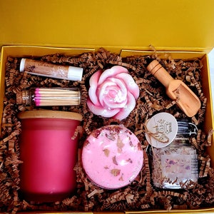 May include: A yellow gift box filled with pink bath and body products. The box contains a pink rose-shaped soap, a pink bath bomb, a jar of bath salts, a pink candle, a tube of lip balm, and a small jar of matches.