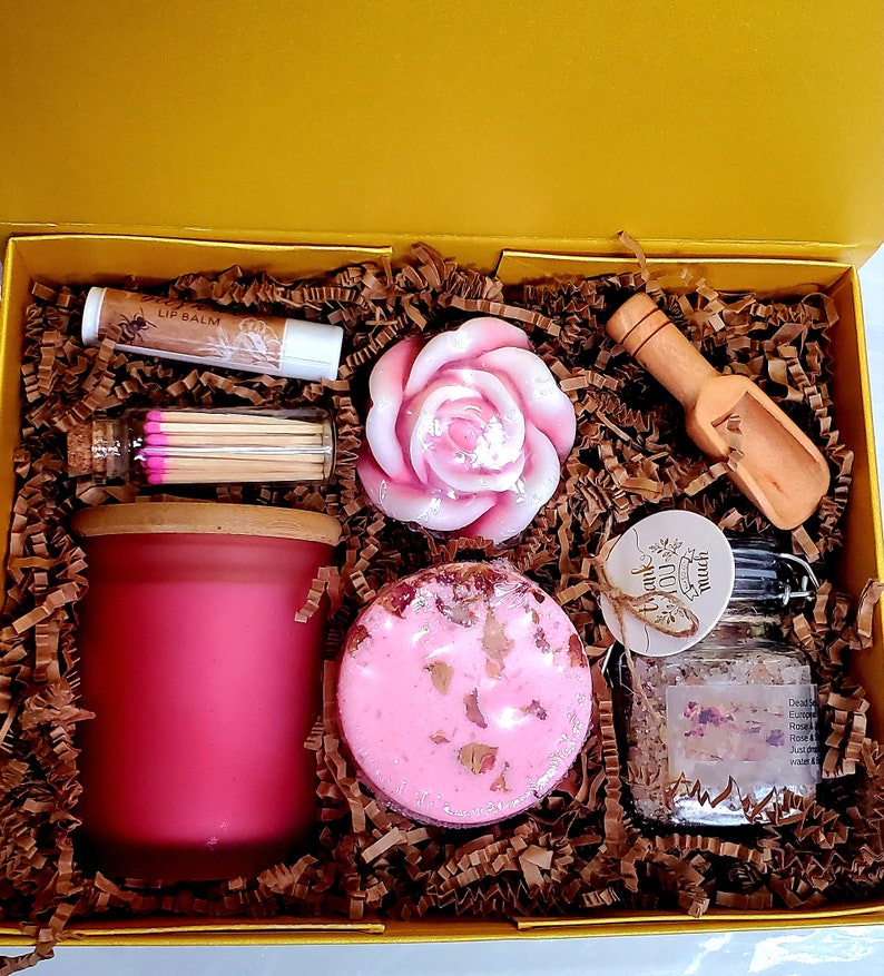 May include: A yellow gift box filled with pink bath and body products. The box contains a pink rose-shaped soap, a pink bath bomb, a jar of bath salts, a pink candle, a tube of lip balm, and a small jar of matches.