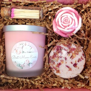 May include: A pink gift box filled with brown shredded paper. The box contains a pink candle with a label that says "Best Mom Ever", a pink rose-shaped soap, and a pink bath bomb with rose petals.