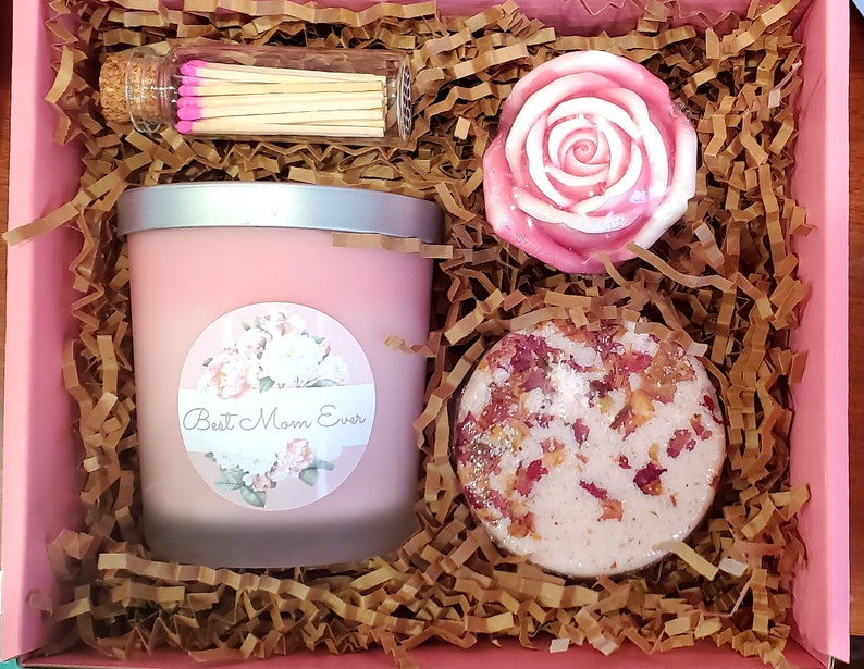 May include: A pink gift box filled with brown shredded paper. The box contains a pink candle with a label that says "Best Mom Ever", a pink rose-shaped soap, and a pink bath bomb with rose petals.