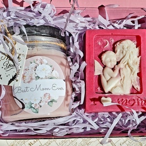 May include: A pink gift box filled with purple shredded paper contains a pink candle with a floral design and the text "Best Mom Ever" and a pink soap with a white sculpted angel and the text "I love Mom".