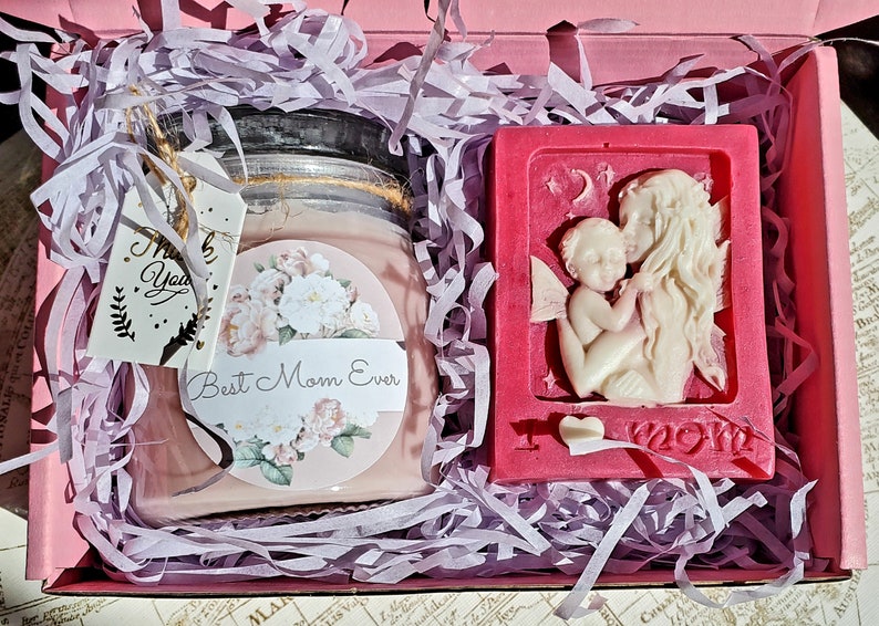 May include: A pink gift box filled with purple shredded paper contains a pink candle with a floral design and the text "Best Mom Ever" and a pink soap with a white sculpted angel and the text "I love Mom".