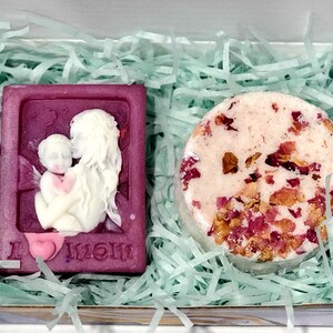May include: A gift box filled with green shredded paper containing a purple soap bar with a white relief image of a mother and child and the text "I love Mom" and a round bath bomb with pink and white swirls and dried rose petals.