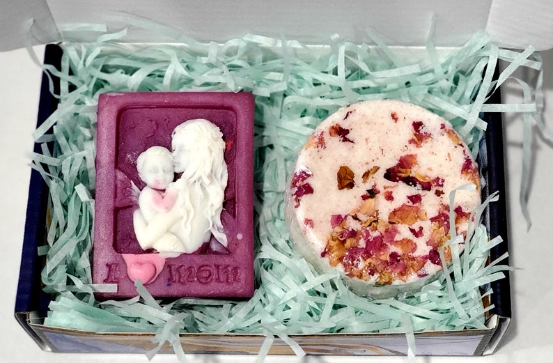May include: A gift box filled with green shredded paper containing a purple soap bar with a white relief image of a mother and child and the text "I love Mom" and a round bath bomb with pink and white swirls and dried rose petals.