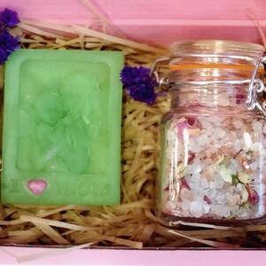 May include: A pink gift box filled with brown packing material. The box contains a green bar of soap with a pink heart and the words "I Love You" and a glass jar filled with pink bath salts and dried flower petals.