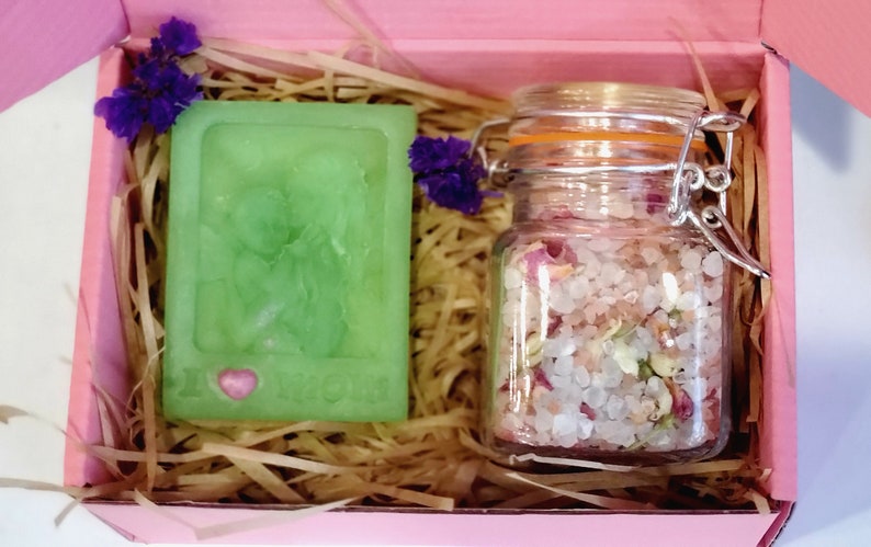 May include: A pink gift box filled with brown packing material. The box contains a green bar of soap with a pink heart and the words "I Love You" and a glass jar filled with pink bath salts and dried flower petals.