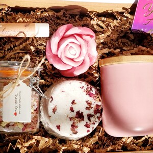 May include: A pink and white self-care gift box with a rose-shaped soap, bath bomb, bath salts, lip balm, and a pink tumbler. The box is filled with brown crinkle paper and includes a card that says "You Are Awesome!"