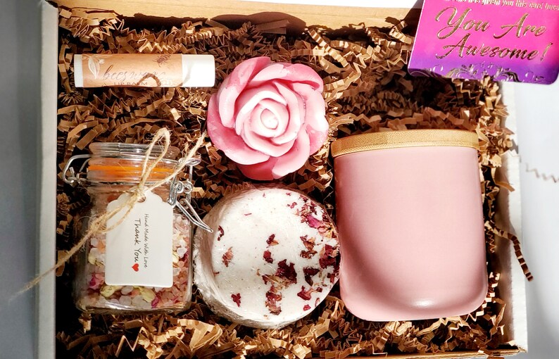 May include: A pink and white self-care gift box with a rose-shaped soap, bath bomb, bath salts, lip balm, and a pink tumbler. The box is filled with brown crinkle paper and includes a card that says "You Are Awesome!"