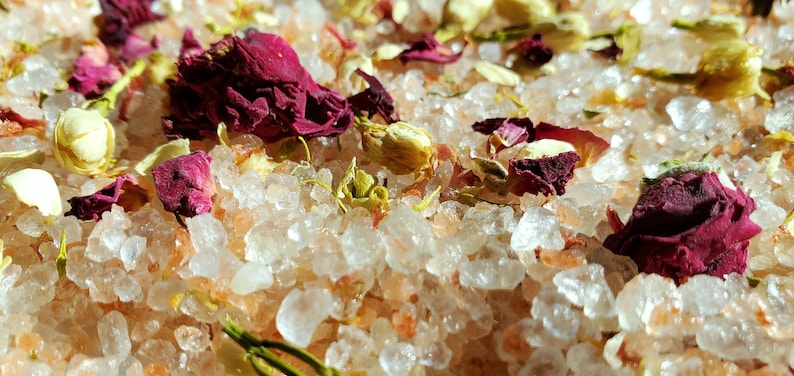 May include: Close-up of pink Himalayan salt bath soak with dried rose petals and white jasmine flowers.