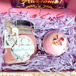 May include: A pink gift box filled with lavender-colored shredded paper. The box contains a pink bath bomb with dried rose petals and a pink candle with a floral design and the text "Best Mom Ever".
