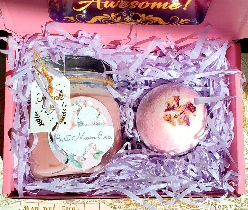 May include: A pink gift box filled with lavender-colored shredded paper. The box contains a pink bath bomb with dried rose petals and a pink candle with a floral design and the text "Best Mom Ever".