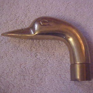 May include: A brass duck head, likely a decorative element or handle. The duck's head is angled, with a detailed beak and eye. The brass has a polished, reflective surface, suggesting it is a vintage or antique item.