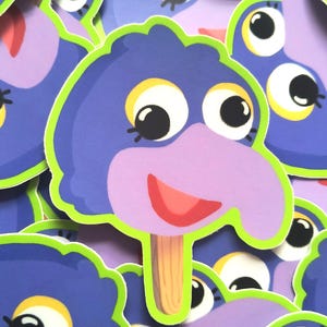 Puede incluir: Una pegatina troquelada con forma de personaje morado parecido a un pájaro sobre un palo de helado de madera. El personaje tiene ojos blancos grandes con pupilas negras, párpados amarillos y un pico morado claro. Borde verde lima brillante.