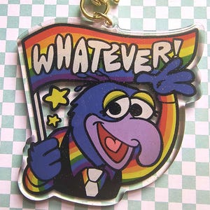 May include: A clear acrylic keychain featuring a purple character with a large nose, wearing a black vest and white tie, holding a rainbow flag with the text WHATEVER! in white letters. The design includes yellow stars and a gold star-shaped clasp.