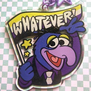 May include: A clear acrylic keychain featuring a purple character with a large nose, wearing a dark suit and tie, holding a flag with the text WHATEVER! in yellow, white, and purple. The design includes glitter accents and a purple star-shaped clasp.