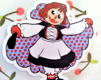 May include: A red-haired rag doll with a black and white dress and a red polka dot cape. The doll is smiling and has large eyes.