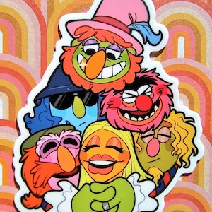 May include: A group of colourful cartoon characters, including a green character with a pink hat, a blue character with sunglasses, a pink character with a red nose, a yellow character with long blonde hair, and a green character with brown hair, are all hugging each other.