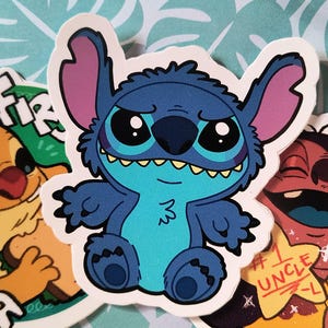 Könnte beinhalten: Eine Sammlung farbenfroher Aufkleber mit Cartoon-Charakteren. Der zentrale Aufkleber zeigt einen blauen Außerirdischen mit großen Augen und einem schelmischen Gesichtsausdruck. Weitere Aufkleber zeigen ein gelbes Wesen mit dem Text "Fir" und eine rote Figur mit dem Text "#1 Uncle L".