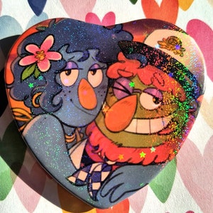 Op de afbeelding: Een hartvormige knoop met een glinsterende afwerking met twee cartoonfiguren. Het personage links heeft blauw haar, een roze bloem en een blauw en geel outfit. Het personage rechts heeft groene huid, rood haar en een zwart-wit geruit overhemd.