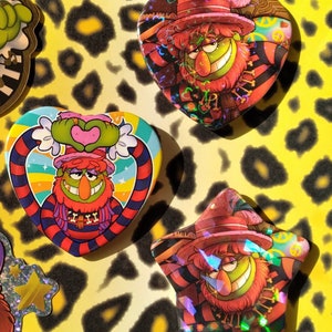 May include: Three heart-shaped and one star-shaped buttons featuring a cartoon character with red hair, a green face, and a striped shirt. The character is wearing a hat and has a large red beard. The buttons have a holographic finish.