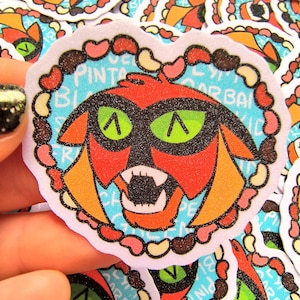 Op de afbeelding: Een hartvormige sticker met een cartoonfiguur met rode vacht, zwarte ogen en een groen masker. Het figuur is omringd door een rand van roze, bruine en witte harten en de tekst "Love, Barbarian Princess".