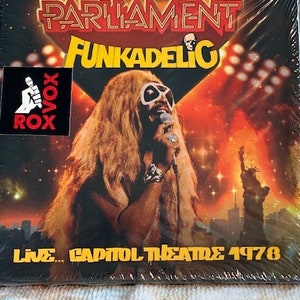 May include: A black and white sticker with the text "Vox Rox" is on the front of a black CD case. The case has a red and yellow image of a man with long blonde hair and sunglasses. The text "Parliament Funkadelic Live... Capitol Theatre 1978" is on the front of the case.