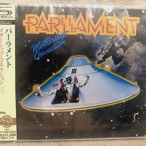 May include: A CD case featuring the album "Mothership Connection" by Parliament. The cover art depicts a silver spaceship with a figure in a spacesuit. The band's name is in orange block letters above the album title.