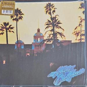 May include: Eagles "Hotel California" vinyl record album cover. The album art features a building with a domed roof, palm trees, and the title in a stylized font. The color palette includes shades of yellow, blue, and brown.
