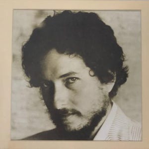 May include: Black and white portrait of a man with dark, curly hair and a beard. The subject is looking towards the viewer. The image is framed in a light beige border.