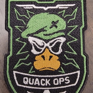 May include: A black and green embroidered patch with the text "Call of Ducky" and "Quack Ops". The patch features a cartoon duck wearing a green beret and sunglasses.