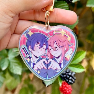 Puede incluir: Un llavero de acrílico en forma de corazón con una colorida ilustración estilo anime de dos personajes con cabello azul oscuro y rosa formando un corazón con sus manos, unido a un cierre de mosquetón de metal dorado.