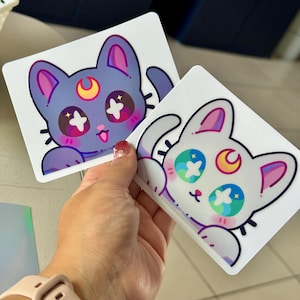Könnte beinhalten: Zwei Aufkleber mit Cartoon-Katzen-Designs. Eine Katze ist lila mit einem gelben Halbmond und sternförmigen Augen. Die andere Katze ist weiß mit einem gelben Halbmond und sternförmigen Augen. Beide Aufkleber haben rosa Akzente.