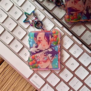 Puede incluir: Un amuleto iridiscente de colores con dos personajes de estilo anime con flores y estrellas. El amuleto está unido a un llavero plateado.