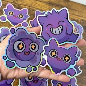 Puede incluir: Una colección de pegatinas holográficas con criaturas moradas de dibujos animados con diversas expresiones. Las pegatinas tienen un borde brillante e iridiscente y se sostienen en una mano. Las criaturas tienen ojos grandes y caras sonrientes.